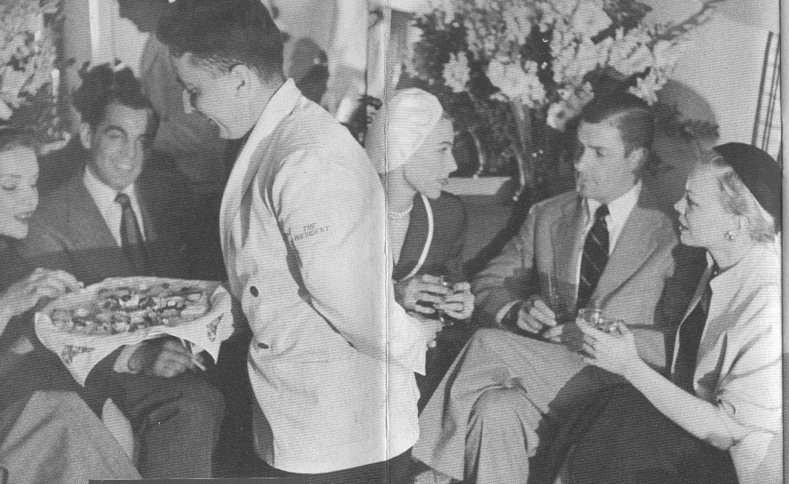 1950s A steward in white serving jacket offers snacks to customers in the lower deck lounge of a Pan Am Boeing 377 Stratocruiser.